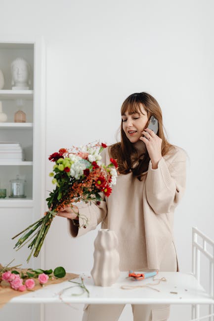 A young woman with long, wavy brown hair and light skin is standing at a white table in a bright, modern room with minimalist decor. She is holding a large, colorful bouquet of fresh flowers in her le