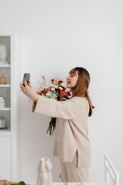 A woman with long, light brown hair and fair skin is holding a vibrant bouquet of fresh flowers in her left arm, while taking a selfie with her right hand. The bouquet features a mix of red, white, an