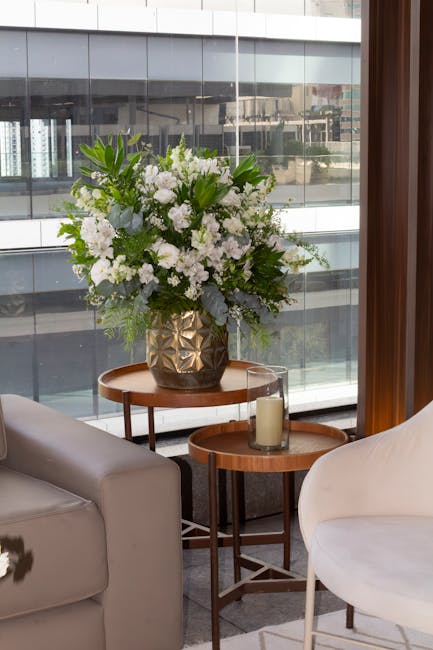 A tall, lush flower arrangement featuring white flowers such as roses, lilies, and hydrangeas, complemented by various green foliage, displayed in a decorative gold geometric-patterned vase. The bouqu
