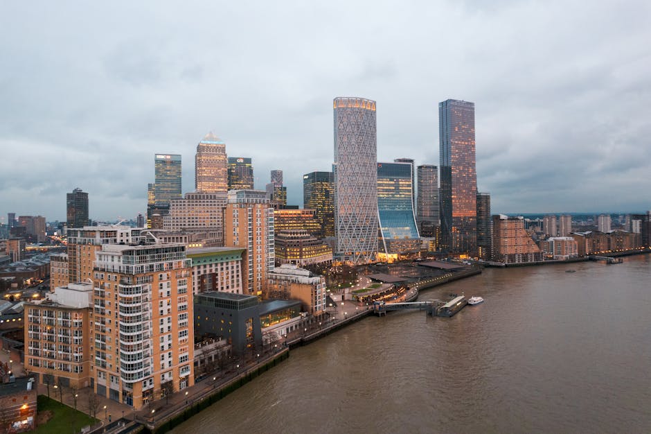 A panoramic view of the Canary Wharf district in London during the evening, featuring a cluster of modern skyscrapers with glass facades reflecting the cloudy sky. The buildings vary in height and des